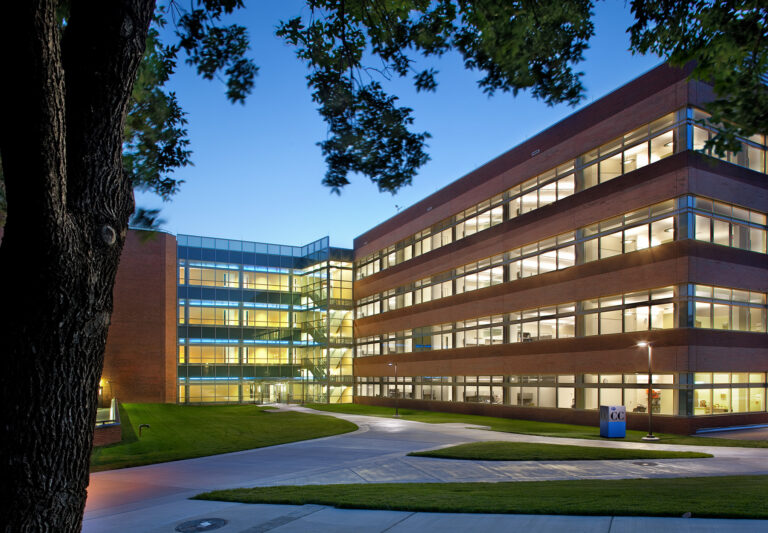 Exterior of a multi-story brick office or academic building at dusk with illuminated windows and green landscaping